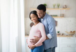 Pregnant black woman and her happy husband hugging next to window at home
